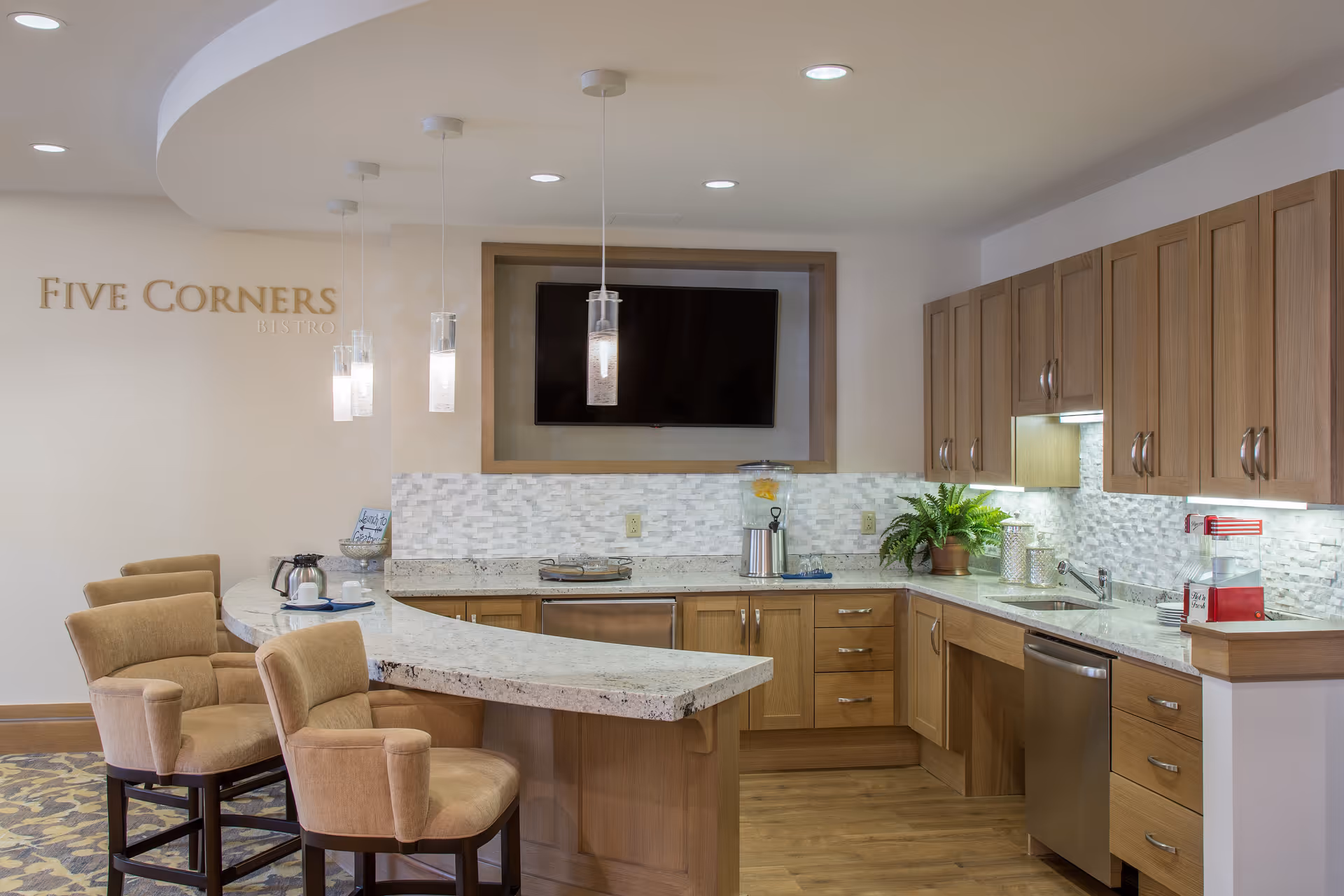 Bright modern kitchen and bistro area with a curved marble-topped counter, upholstered bar chairs, wooden cabinets, and a wall-mounted TV.