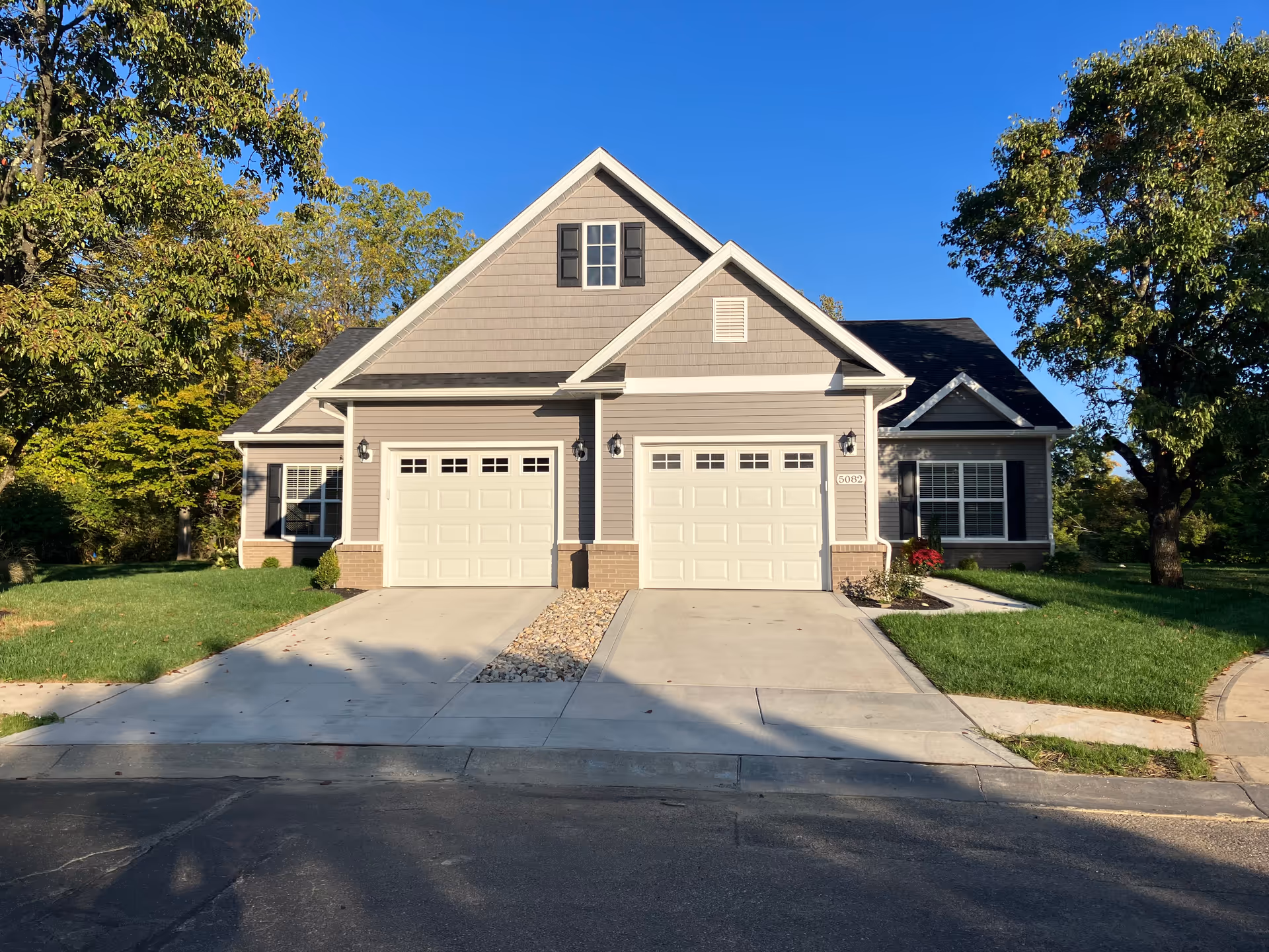 Front view of a duplex-style house with two garage doors, a shared driveway, lawn and trees under a clear blue sky.