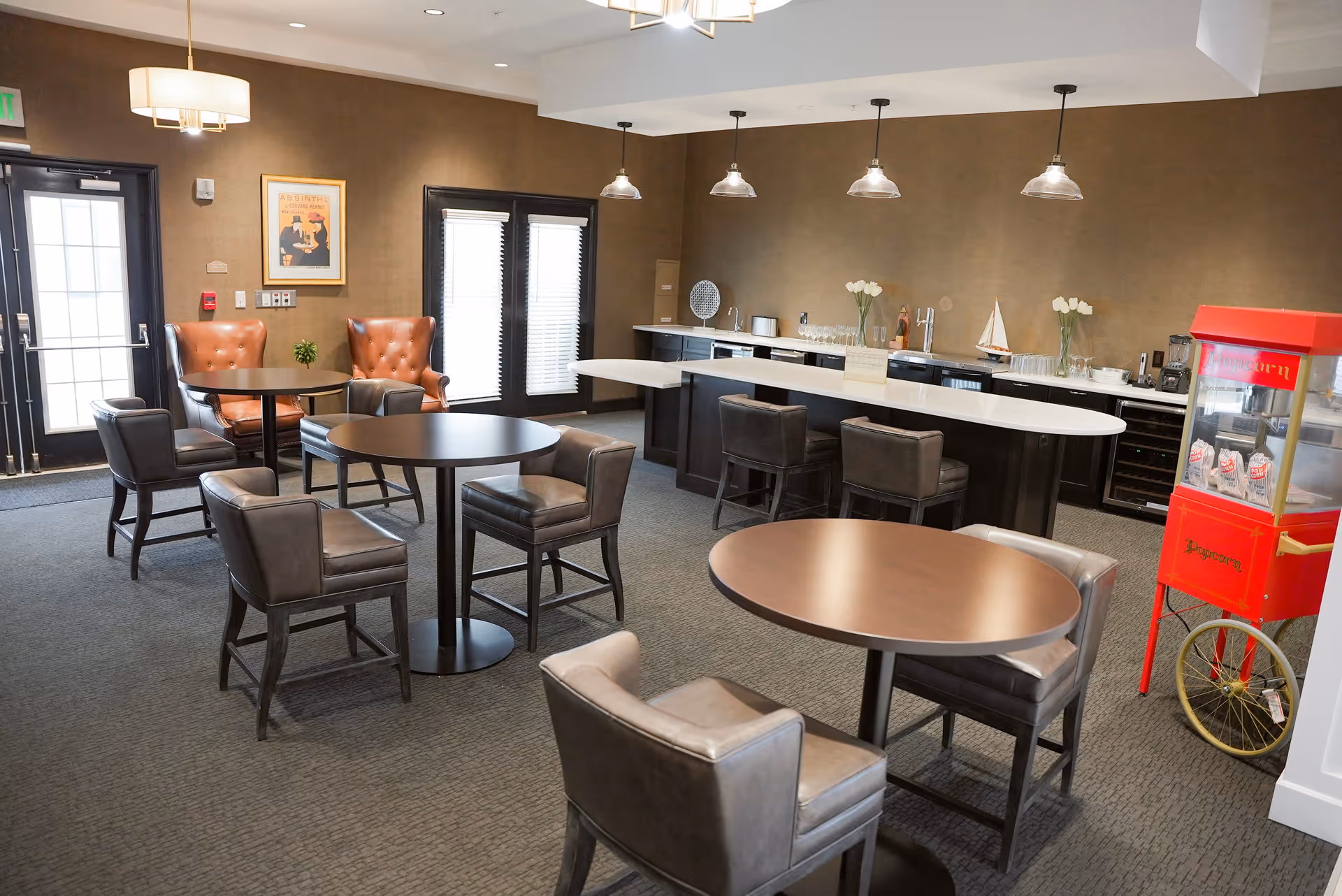 A cozy common area with several round tables and leather chairs arranged around them. There is a long counter with bar stools, a popcorn machine, and decorative items including vases with white flowers and a small model sailboat. The room has brown walls, carpeted floor, and large windows with blinds.