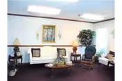 A nursing-home lounge with sofas and armchairs arranged around a central coffee table, lamps, a potted plant, and framed artwork on the wall.
