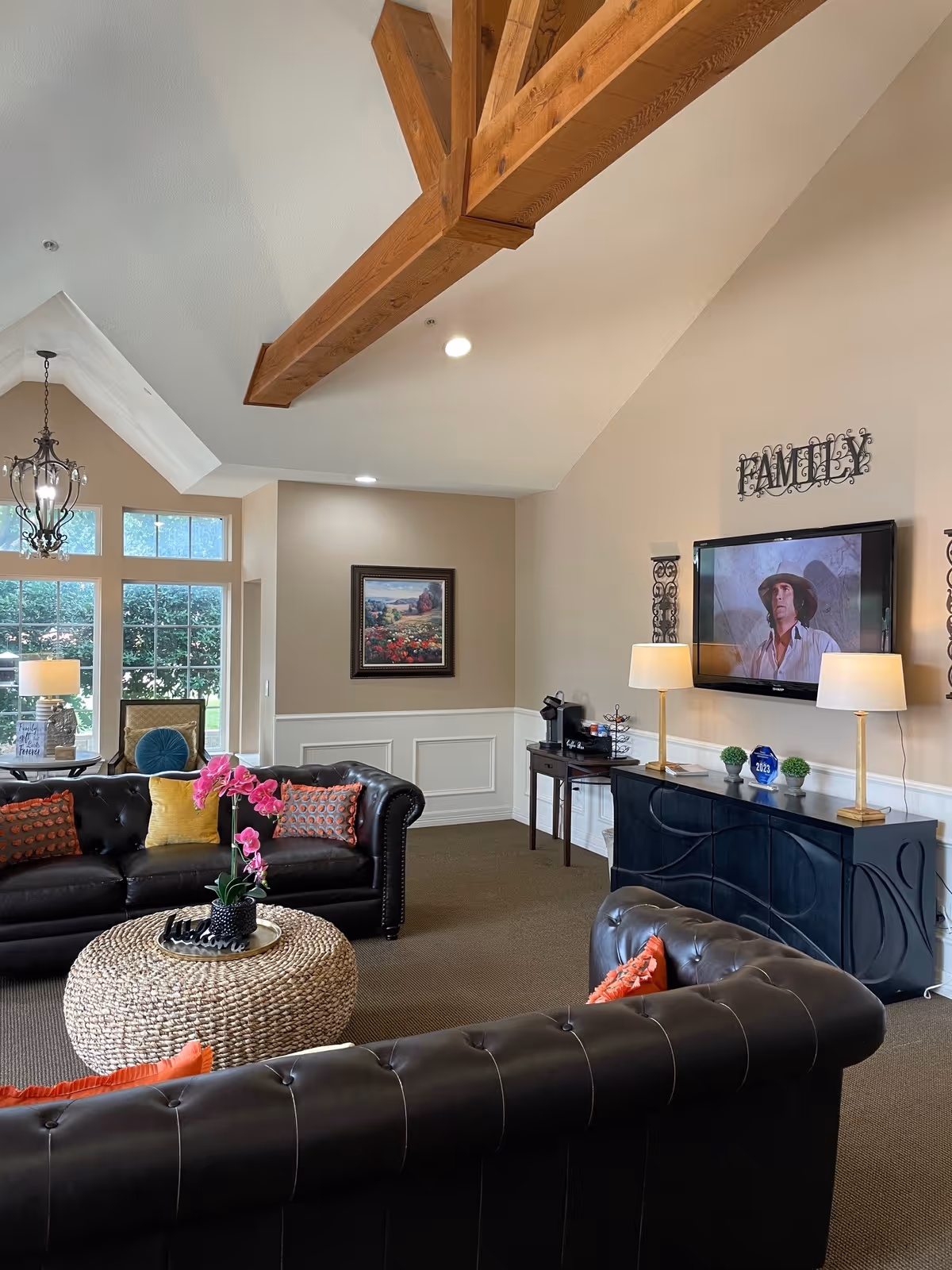 A cozy living room with vaulted ceilings featuring exposed wooden beams. The room has two black leather tufted sofas adorned with colorful pillows, a round woven coffee table with a pink orchid centerpiece, and a large window letting in natural light. On one wall, there is a flat-screen TV mounted above a black console table with two lamps and decorative items. A framed landscape painting hangs on an adjacent wall, and a chandelier is visible near the window.
