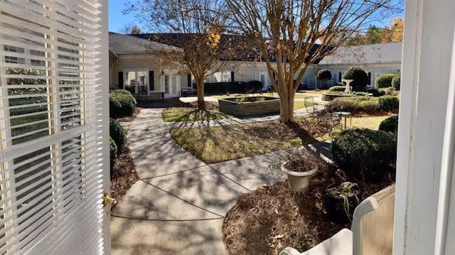 View from an open door looking out onto a courtyard with paved walkways, leafless trees, shrubs, and a raised garden bed in the center, surrounded by a single-story building under a clear blue sky.