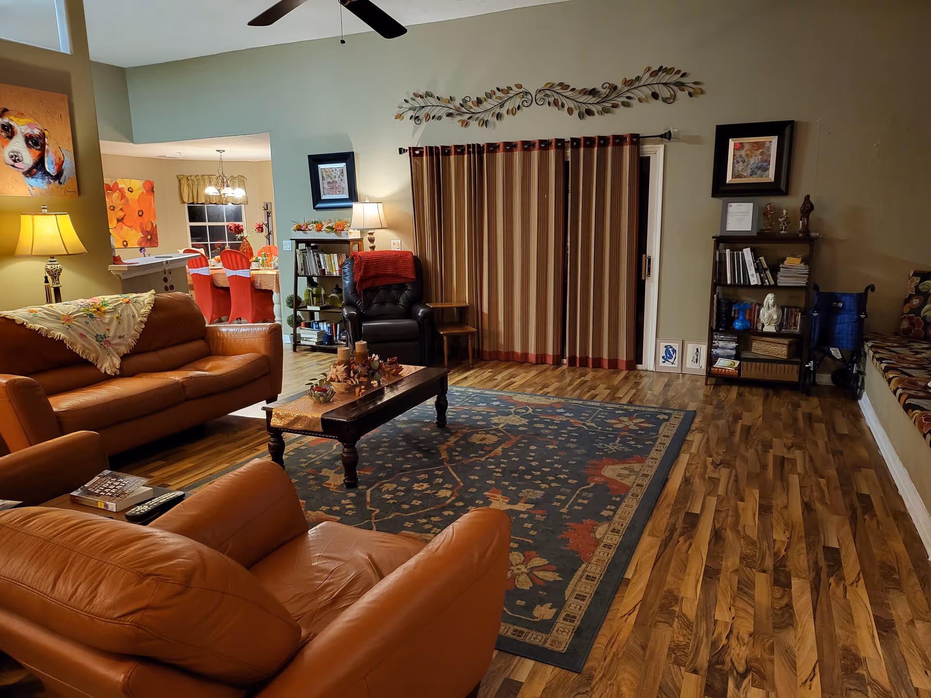 A cozy living room with brown leather sofas and a matching armchair arranged around a wooden coffee table on a patterned rug. The room has wooden flooring, a ceiling fan, and a large window covered with striped curtains. There are bookshelves with books and decorative items, a lamp, and colorful artwork on the walls. In the background, a dining area with a table and red chairs is visible.