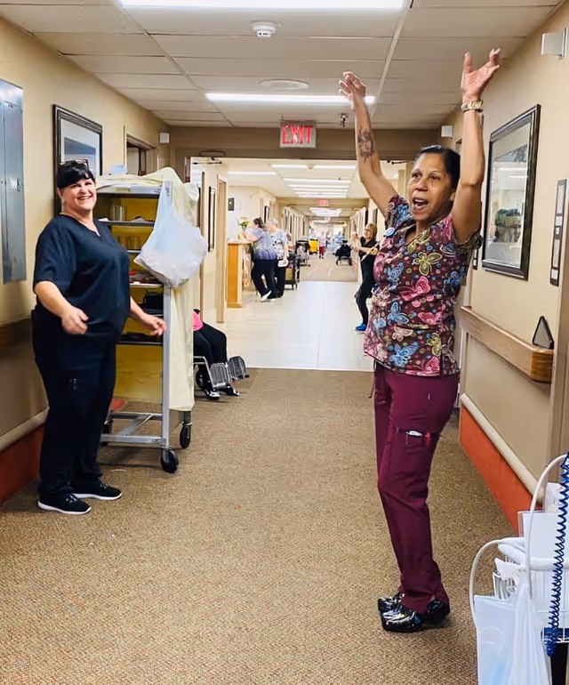 Two women in a hallway of a care facility, one with arms raised and the other smiling, with other people visible further down the corridor. The hallway has beige walls, carpeted floor, and framed pictures on the walls.