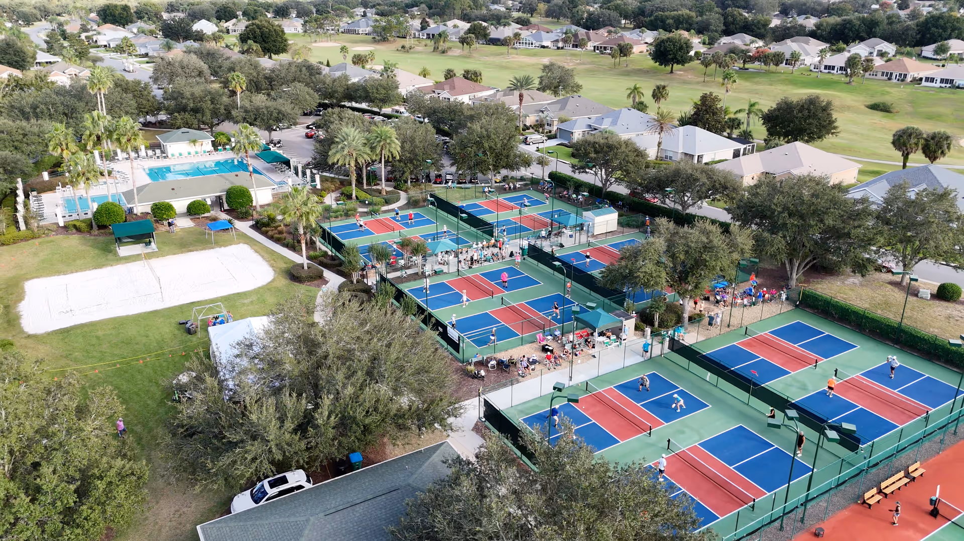 Aerial view of multiple outdoor pickleball/tennis courts, a swimming pool, and surrounding homes in a residential community.