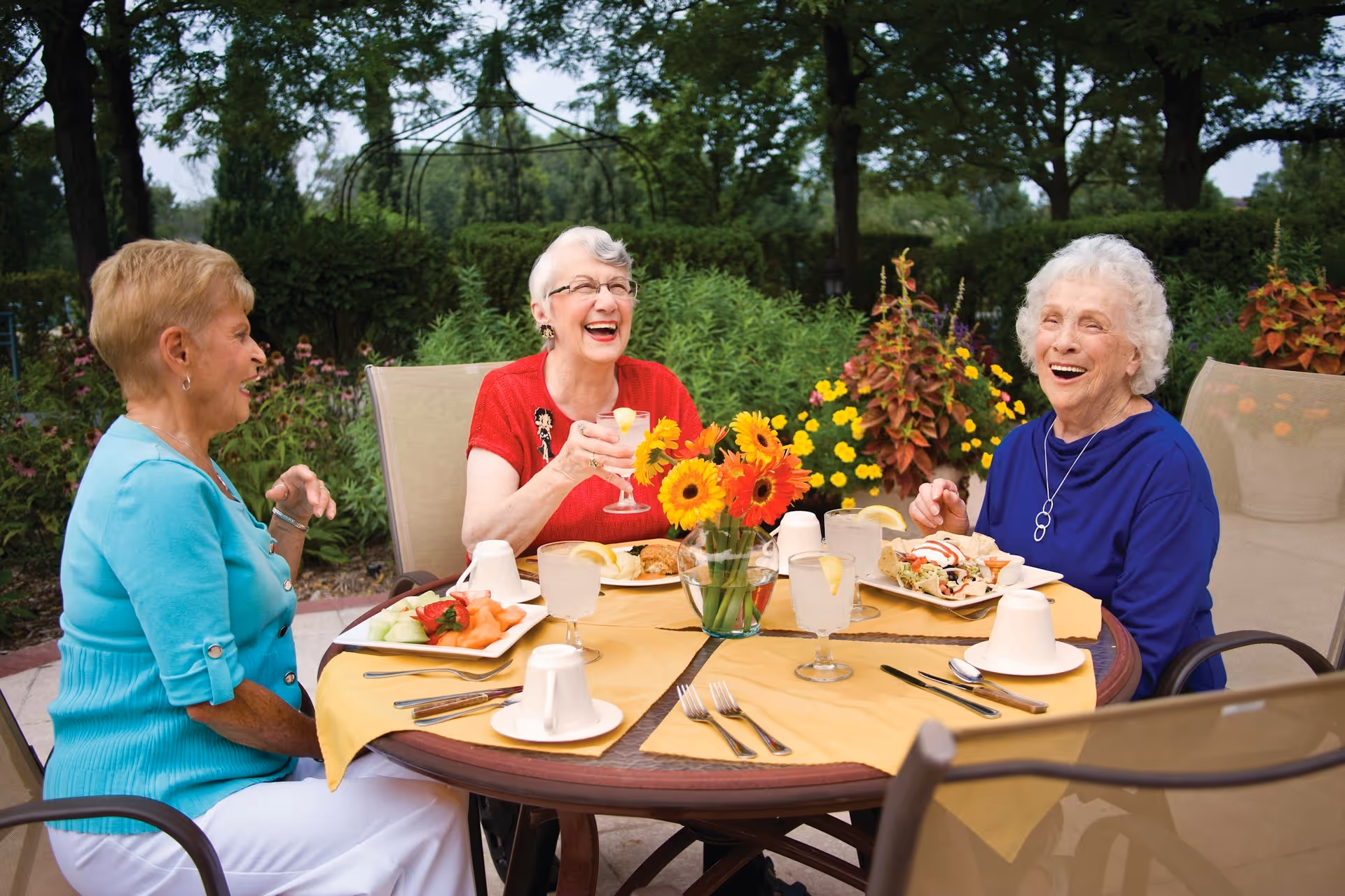 Three older women laugh and enjoy a meal together at a round outdoor patio table decorated with flowers and drinks.