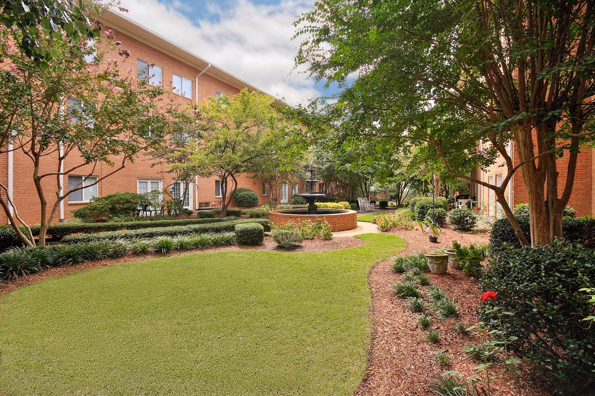 A landscaped outdoor courtyard area with green grass, trees, shrubs, and a central water fountain surrounded by a brick wall. The courtyard is flanked by a three-story brick building with windows and doors leading to the outdoor space. There are also some chairs and a pathway winding through the garden.