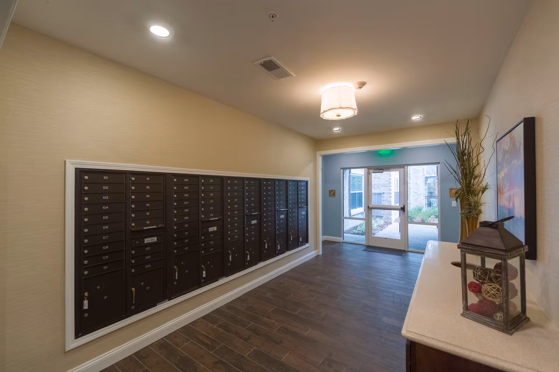Interior hallway with a wall of mailboxes on the left and a console table with decorative items on the right, leading to a glass door exit with a view of the outdoor area.