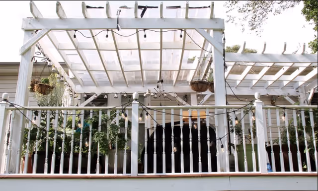 Outdoor patio area with a white wooden pergola and railing, decorated with hanging plants and string lights.