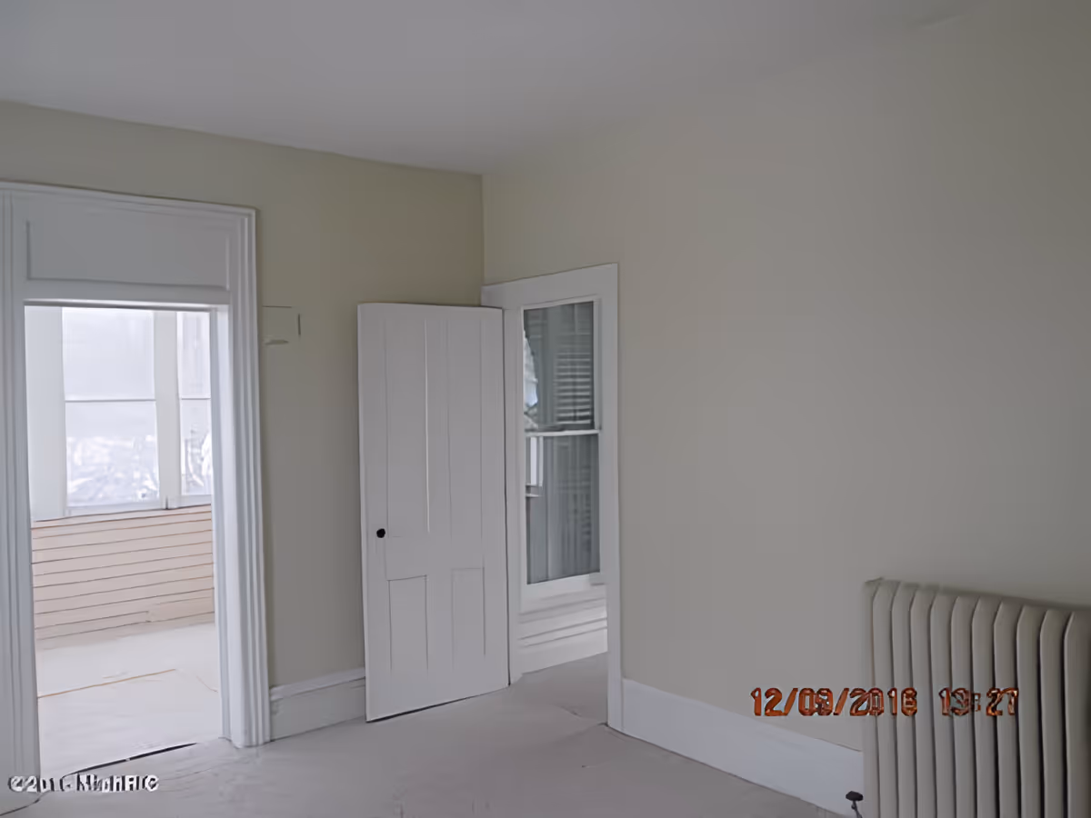 Empty room with beige walls, a white door slightly ajar, a window with white trim, and a radiator on the right side. There is an open doorway leading to a sunroom or enclosed porch with large windows.