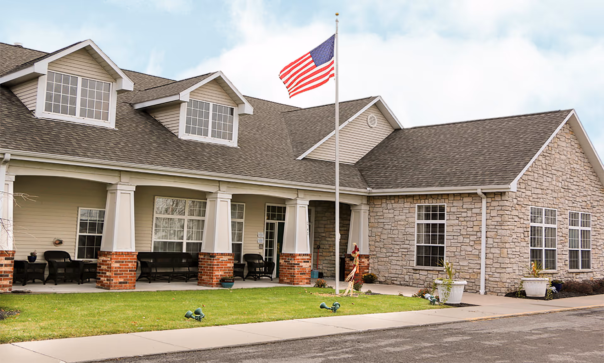 Exterior view of a single-story senior living facility building with a combination of beige siding and stone facade. The building features a covered porch with brick pillars, outdoor seating, and an American flag on a flagpole in front. The sky is partly cloudy.