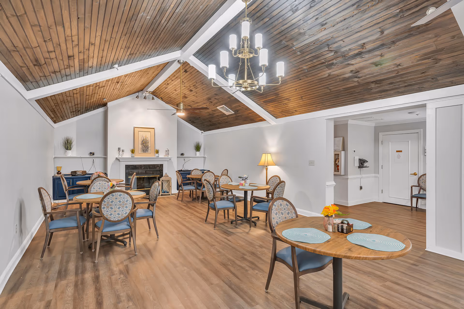A cozy dining room with wooden floors and a vaulted wooden ceiling featuring white beams. The room contains several round tables with patterned chairs, each table set with placemats and small flower arrangements. A chandelier and a ceiling fan hang from the ceiling. There is a fireplace at the far end of the room flanked by built-in shelves with decorative items and plants. A floor lamp is positioned near the wall, and a doorway leads to another area of the facility.
