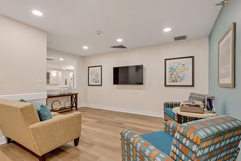 A cozy living room area in an assisted living facility featuring a beige armchair with a blue cushion, two patterned armchairs with blue and orange circular designs, a small round table with a decorative item, a wall-mounted flat-screen TV, and framed artwork on the walls. The room has wood flooring and recessed ceiling lights.