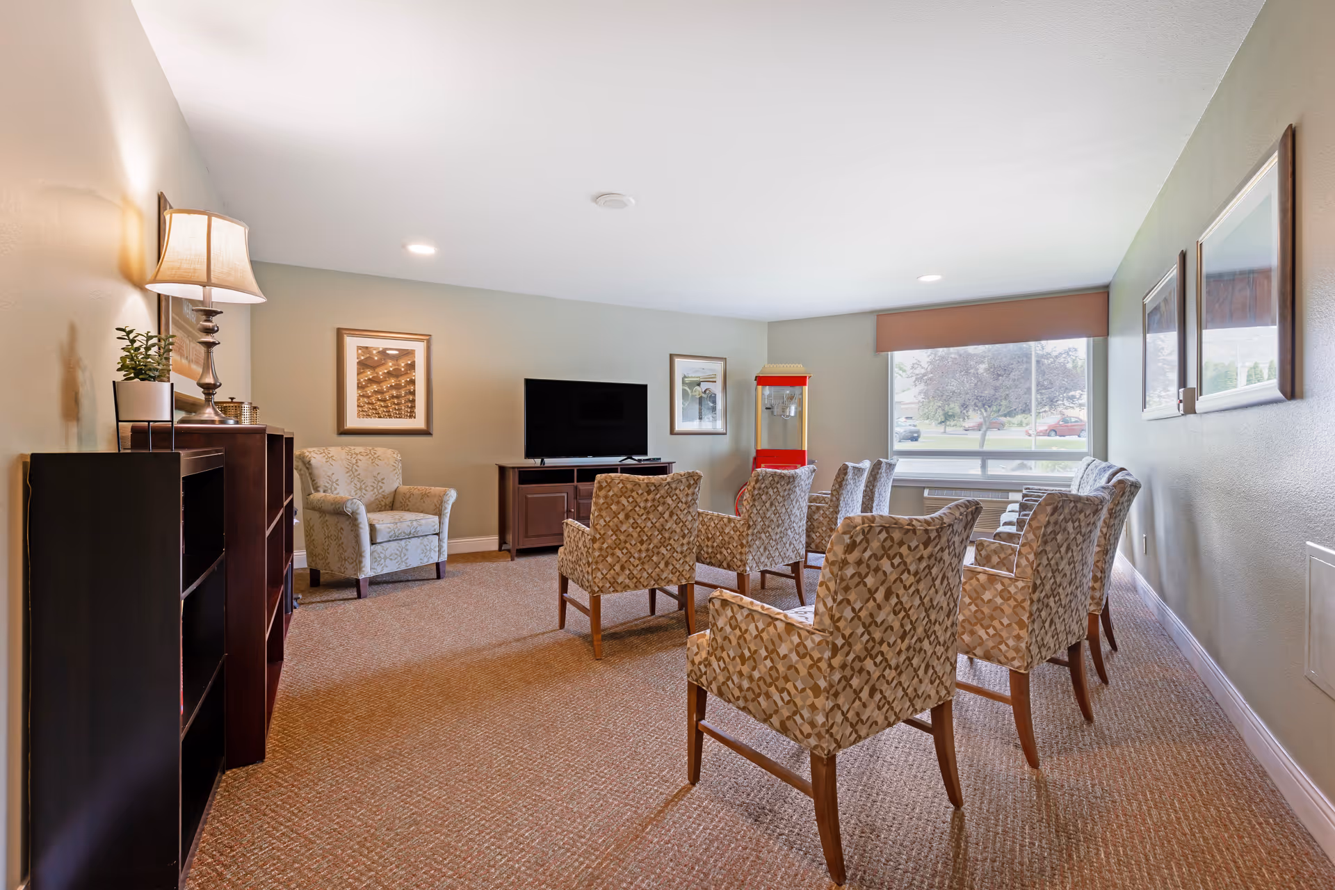 A well-lit living room area with multiple patterned armchairs arranged facing a flat-screen TV on a wooden stand. There is a lamp on a side table, framed artwork on the walls, a popcorn machine near a large window, and a bookshelf on the left side.