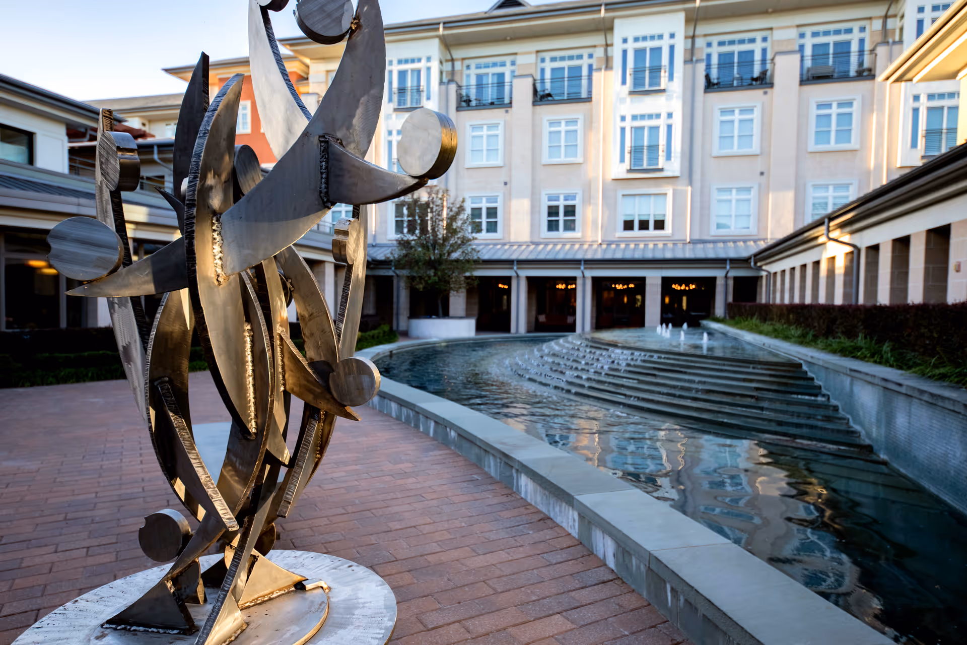 Outdoor courtyard area at The Legacy Willow Bend featuring a modern abstract metal sculpture on a brick pathway next to a tiered water fountain with a multi-story building in the background.