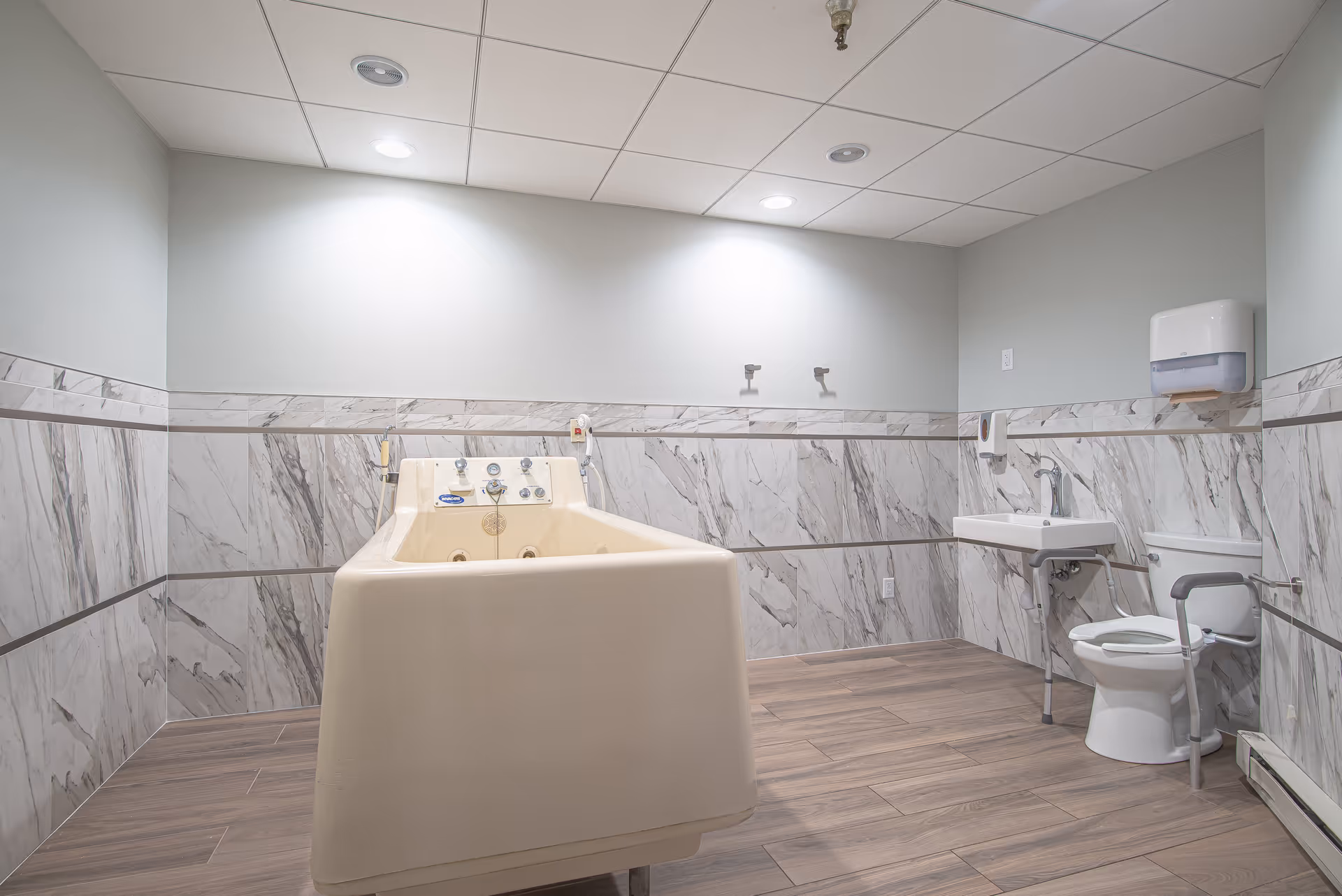 A spacious bathroom with a large beige bathtub in the center, a white toilet with support rails, a wall-mounted sink, and a paper towel dispenser. The walls are partially tiled with a marble pattern, and the floor has wood-like tiles. The ceiling has recessed lighting.