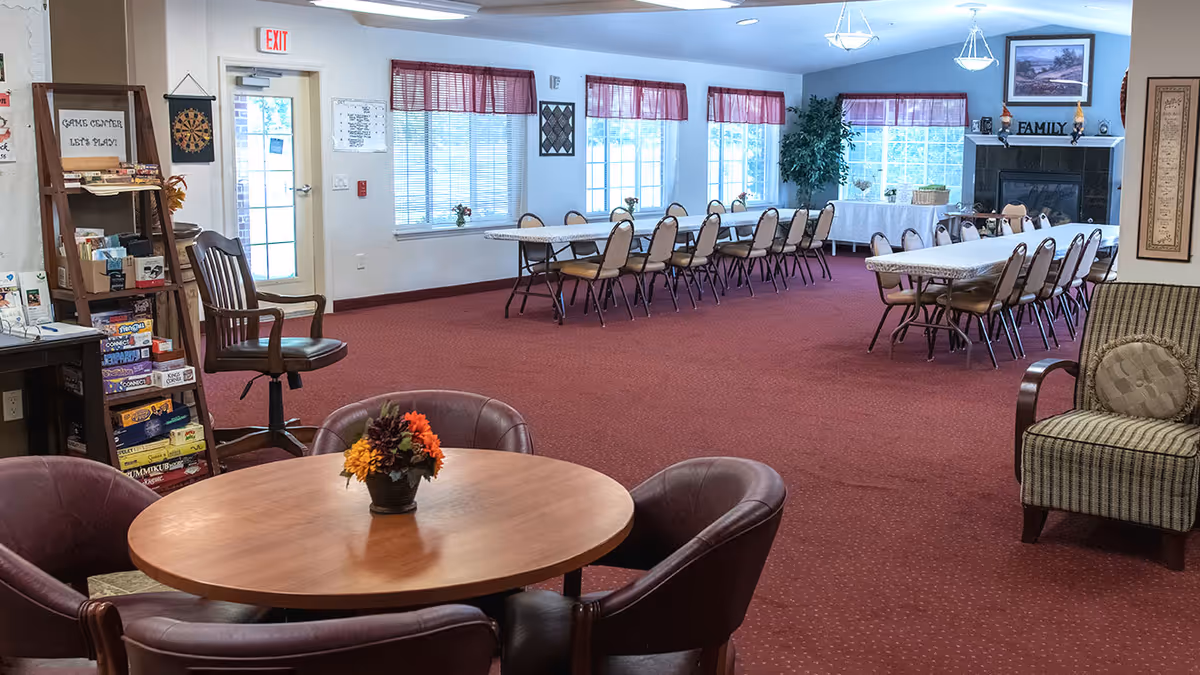A spacious community room with red carpet and multiple tables and chairs arranged for group activities or dining. In the foreground, there is a round wooden table with four leather chairs and a small flower arrangement. In the background, there are two long rectangular tables with chairs, large windows with red valances, a fireplace with a 'FAMILY' sign on the mantel, and a tall green plant. To the left, there is a wooden shelf labeled 'Game Center Let's Play' stocked with board games and a dartboard hanging on the wall near an exit door.