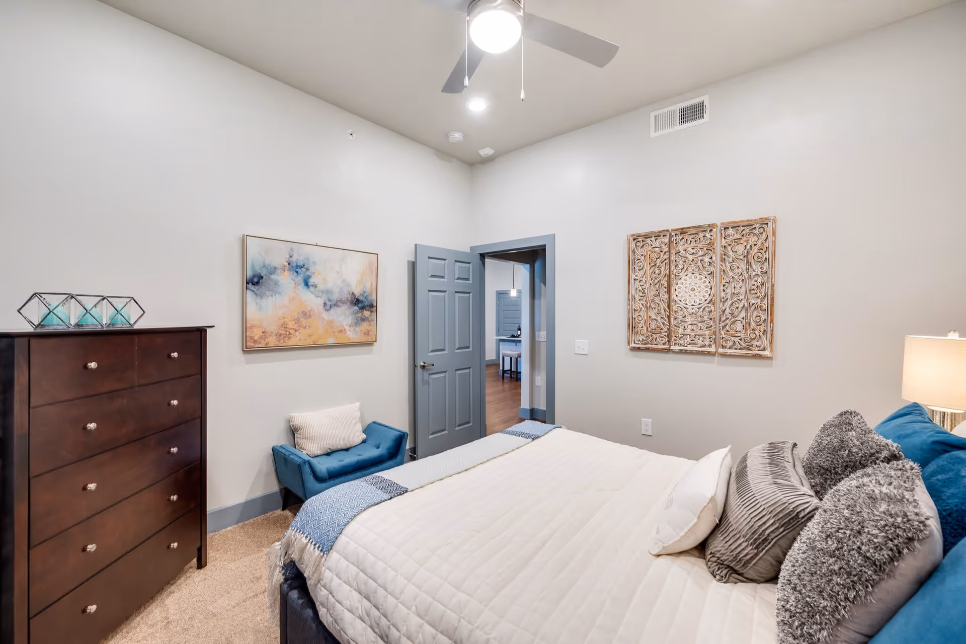 A cozy bedroom in a senior living facility featuring a neatly made bed with multiple pillows and a white quilt. The room has beige walls with two pieces of wall art, a dark wooden dresser with decorative items on top, a small blue upholstered bench with a white pillow, and a ceiling fan with a light. The door is open, showing a glimpse of another room with wooden flooring.