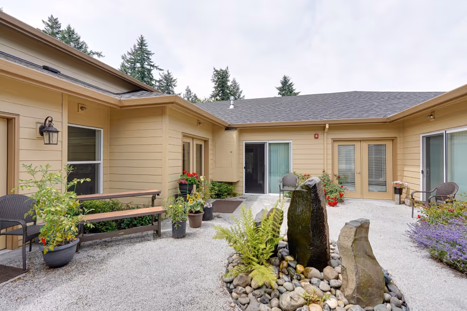 Outdoor courtyard area of a senior care facility with beige siding buildings surrounding a gravel ground. There are potted plants, flowers, and a small rock water feature in the center. Several chairs and a bench are placed around the courtyard, with trees visible in the background under a cloudy sky.