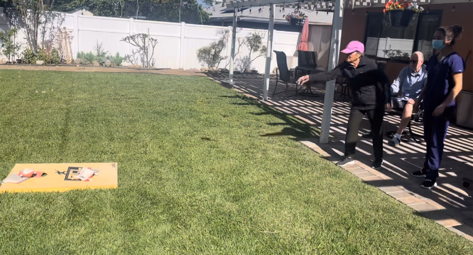 An elderly woman wearing a pink cap is playing a lawn game on a grassy yard, throwing an object towards a yellow board with holes. A man is seated nearby on a patio under a pergola, and a caregiver wearing a mask and navy scrubs stands beside them. The area is enclosed by a white fence with some plants and trees along the perimeter.