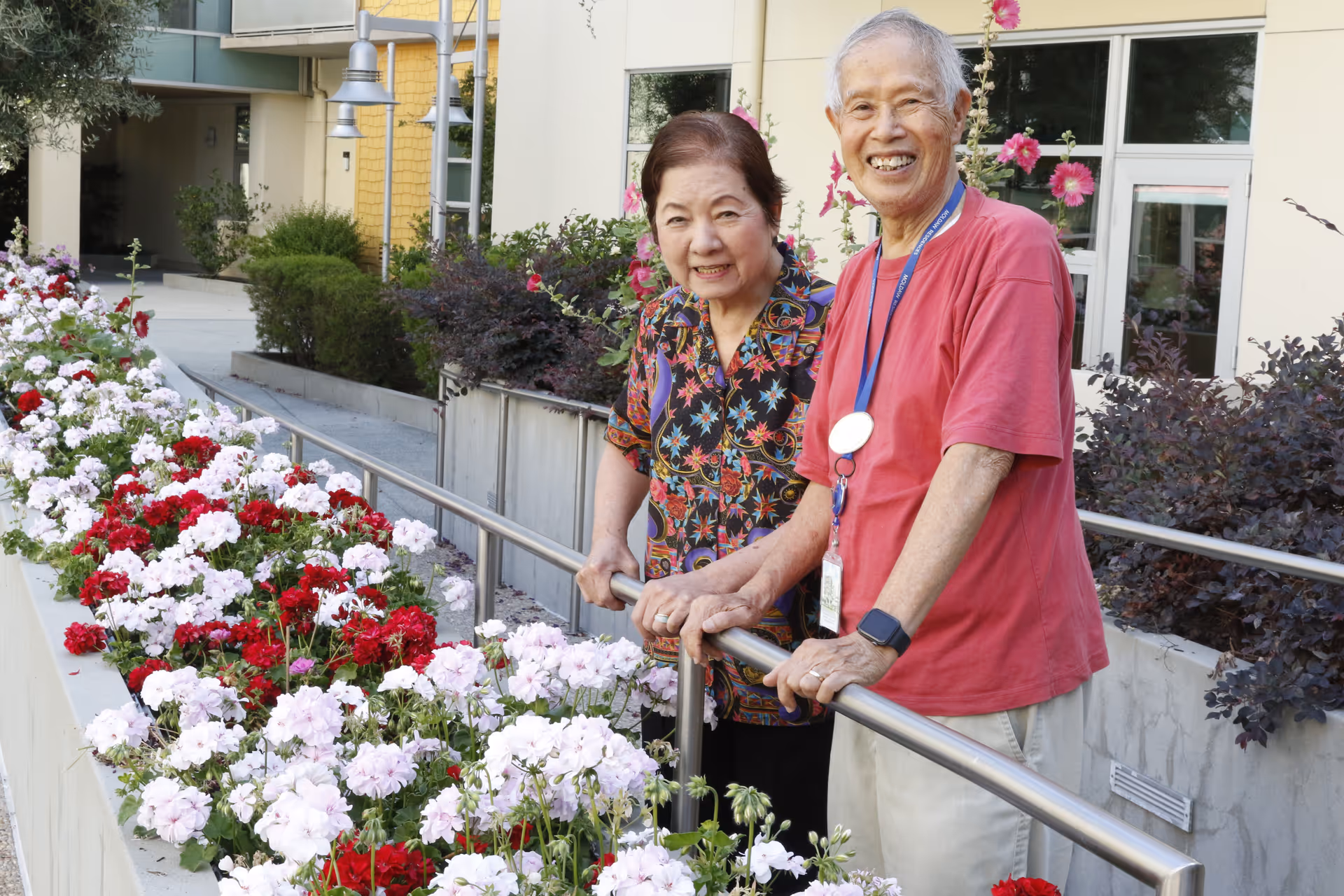 An elderly man and woman standing outdoors next to a flower bed filled with red and white flowers. They are smiling and leaning on a metal railing with a building and greenery in the background.