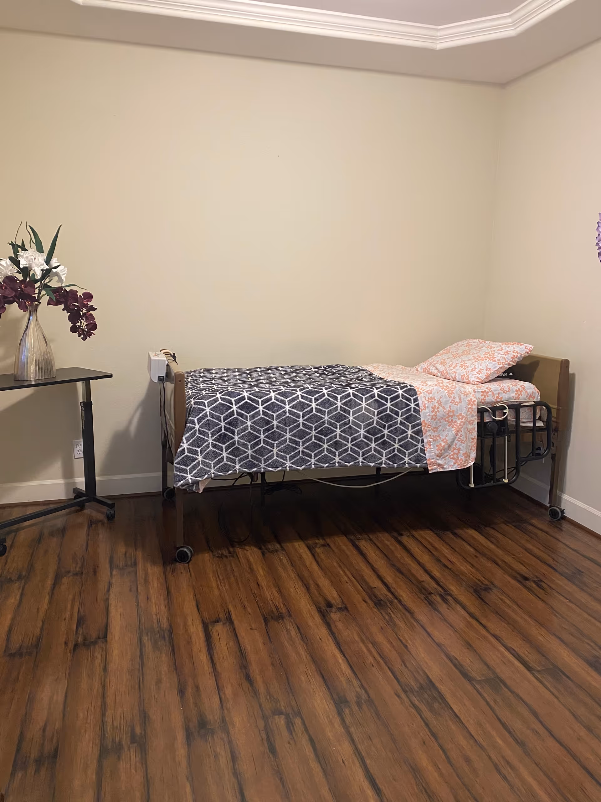 A simple bedroom with a single hospital-style bed covered with a gray and white geometric patterned blanket and a floral pillow and sheet. There is a small table with a vase of flowers on the left side against a beige wall and wooden flooring.
