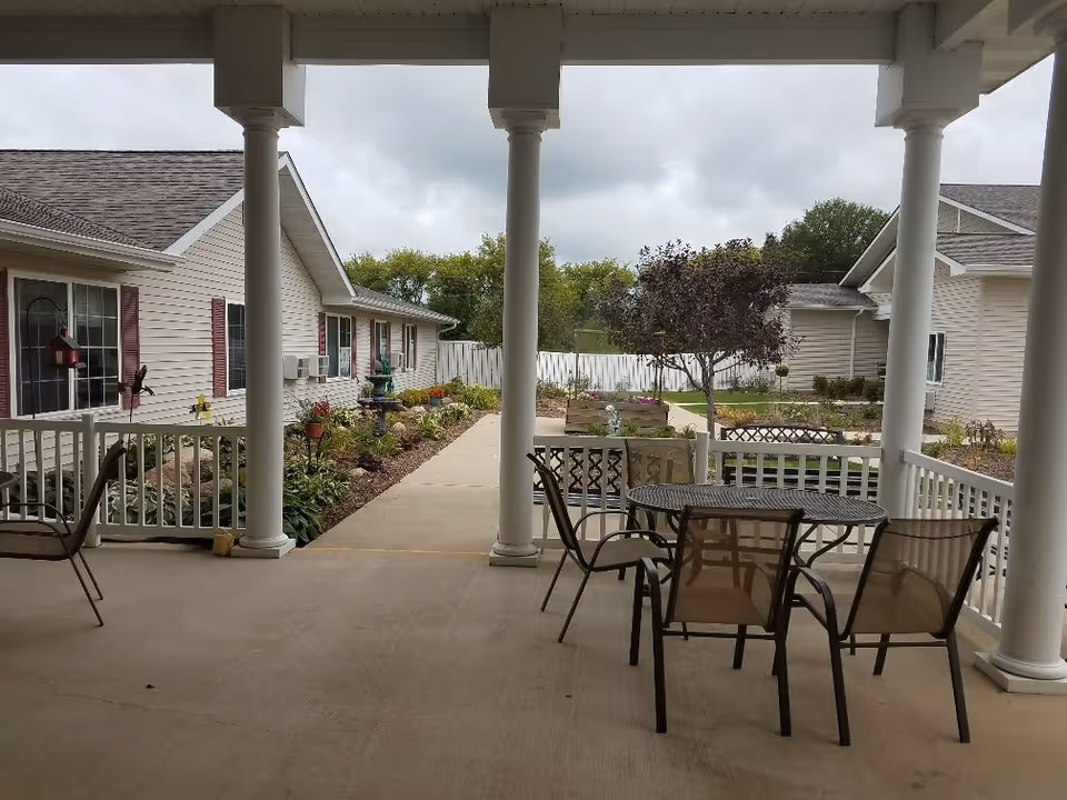 Covered outdoor patio area with a round metal table and four chairs. The patio overlooks a garden path with flower beds, small trees, and shrubs. Two single-story buildings with beige siding and red shutters are visible on either side of the path under a cloudy sky.