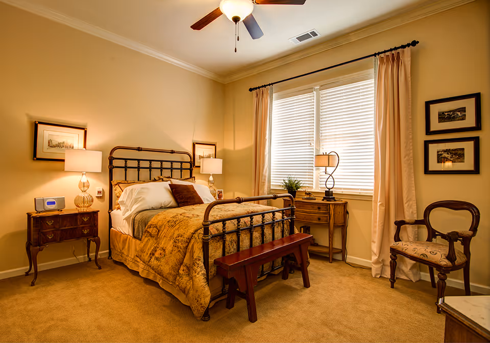 A warmly lit bedroom with a metal framed bed covered in a patterned golden bedspread and white pillows. There are two bedside tables with lamps on either side of the bed, a wooden bench at the foot of the bed, a wooden chair with an upholstered seat, and a small wooden desk with a lamp and a plant near a window with closed blinds and beige curtains.