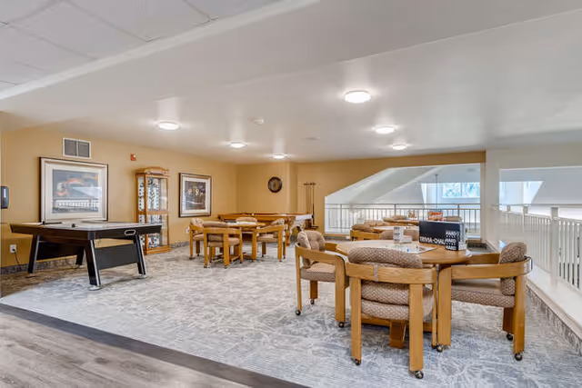 A spacious common area in a senior living community featuring multiple wooden tables and chairs arranged for group activities. There is an air hockey table on the left side, framed artwork on the walls, and a display cabinet. The room has a light-colored carpet and ceiling lights, with a railing overlooking a lower level on the right side.