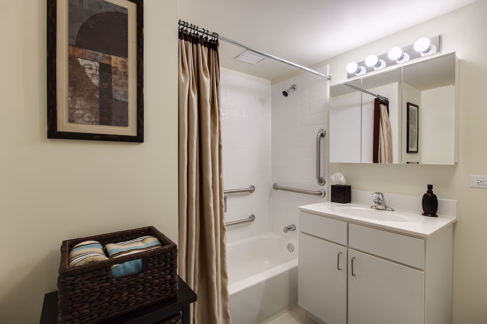 A clean and well-lit bathroom featuring a white bathtub with a beige shower curtain, grab bars installed on the tiled walls for safety, a white vanity with a sink, a tissue box, and a soap dispenser. Above the sink is a large mirror with lights. A framed abstract artwork hangs on the wall next to a basket holding folded towels.