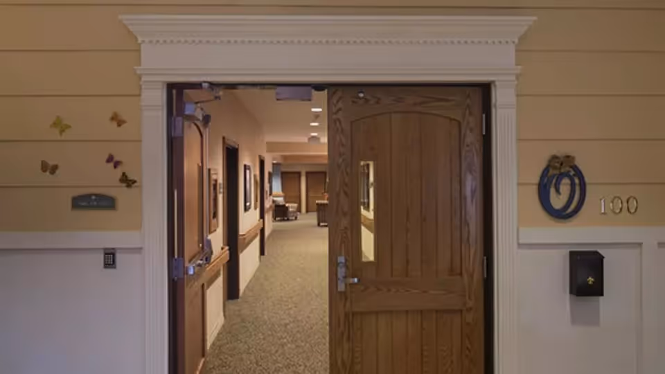 Open wooden doorway leading into a carpeted corridor of a memory care facility with decorative butterflies on the left wall and room number 100 on the right.
