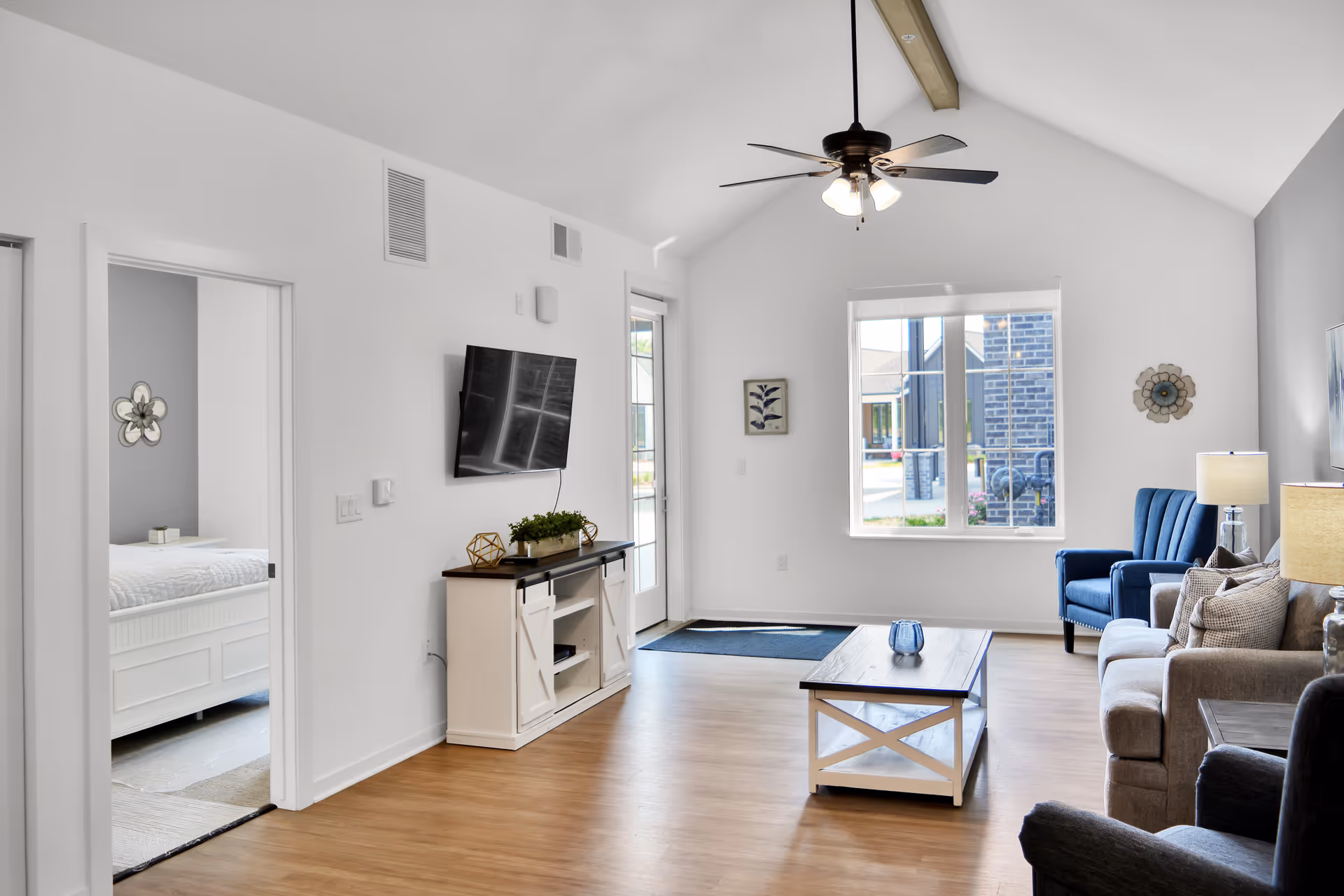 A bright and modern living room with a vaulted ceiling and ceiling fan. The room features a mounted flat-screen TV above a white console table with decorative items, a wooden coffee table, a gray sofa, a blue armchair, and a window showing an exterior view. To the left, an open doorway reveals a bedroom with a white bed and a decorative wall piece.