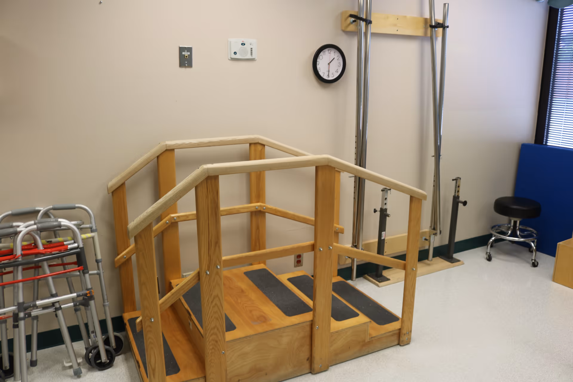 A small indoor physical therapy area with a wooden step platform equipped with handrails, several walkers lined up against the wall, a black stool on wheels, and some metal exercise or support equipment. A clock is mounted on the beige wall above the platform.