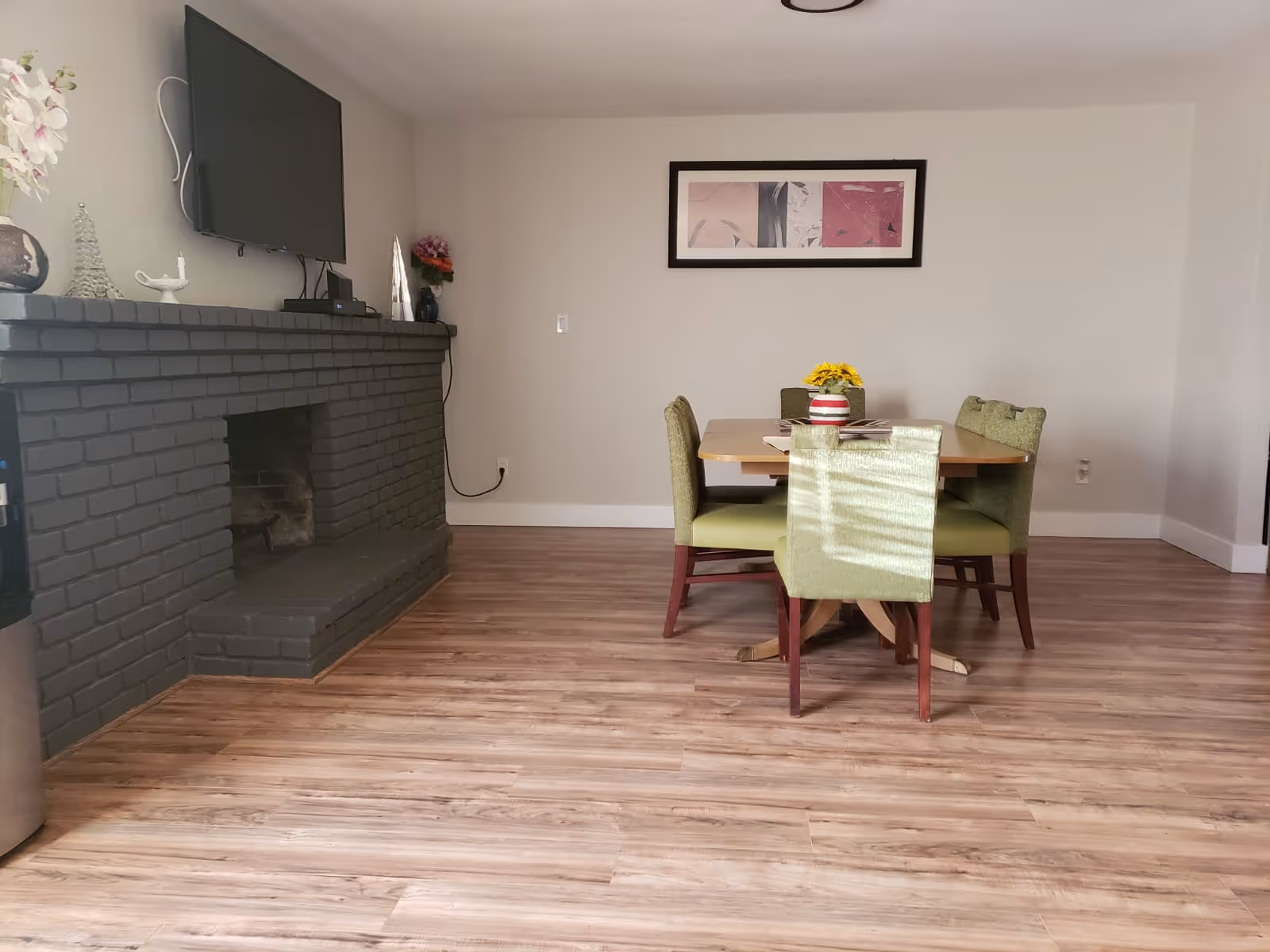 A dining area with a wooden table and four green upholstered chairs. On the table is a vase with yellow flowers and some magazines. To the left is a dark gray brick fireplace with decorative items on the mantel, and a flat-screen TV mounted above it. The floor is light wood, and a framed abstract artwork hangs on the wall behind the table.