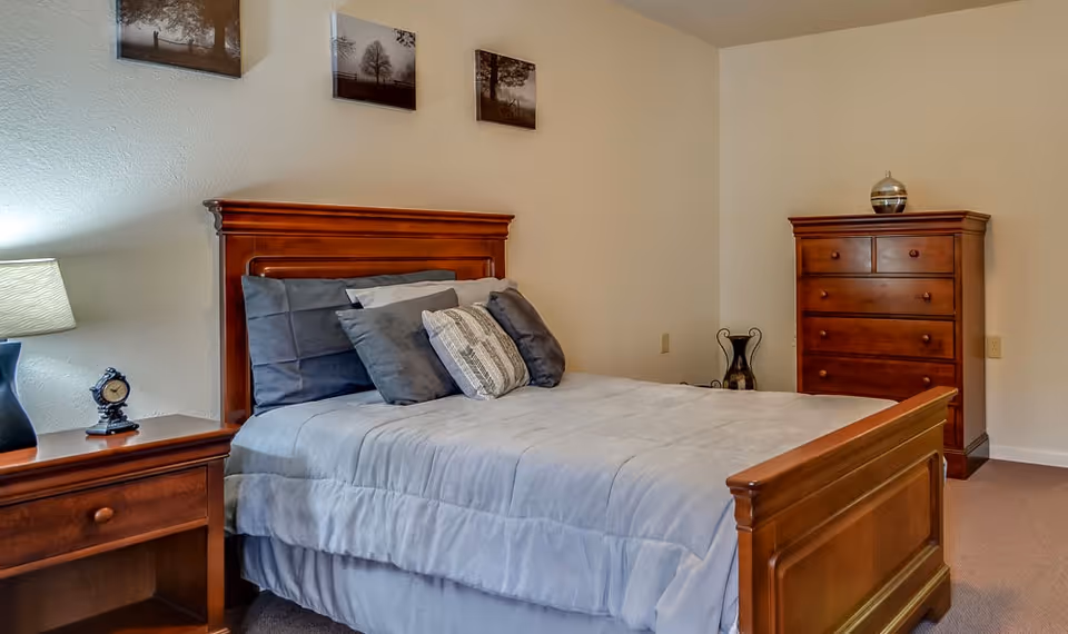 A neatly made bed with gray bedding and multiple pillows in a senior living bedroom. The room features wooden furniture including a nightstand with a lamp and an alarm clock, a dresser with decorative items, and three framed pictures hanging on the wall above the bed.