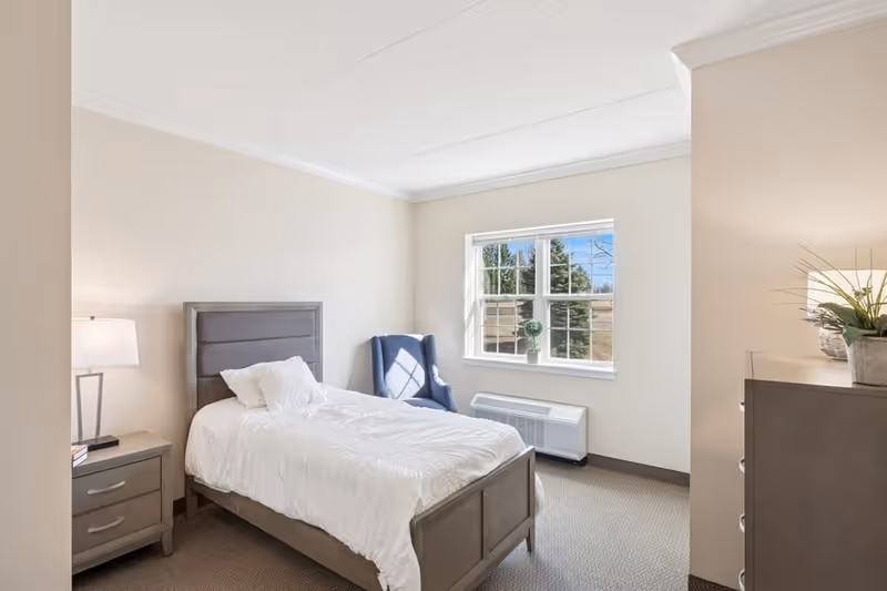 A bright and clean senior living bedroom with a single bed featuring a gray upholstered headboard, white bedding, and a pillow. Next to the bed is a nightstand with a lamp and a book. A blue armchair is positioned near a window that lets in natural light. There is a heater below the window and a dresser with a plant on top on the right side of the room. The walls are painted light beige and the carpet is gray.