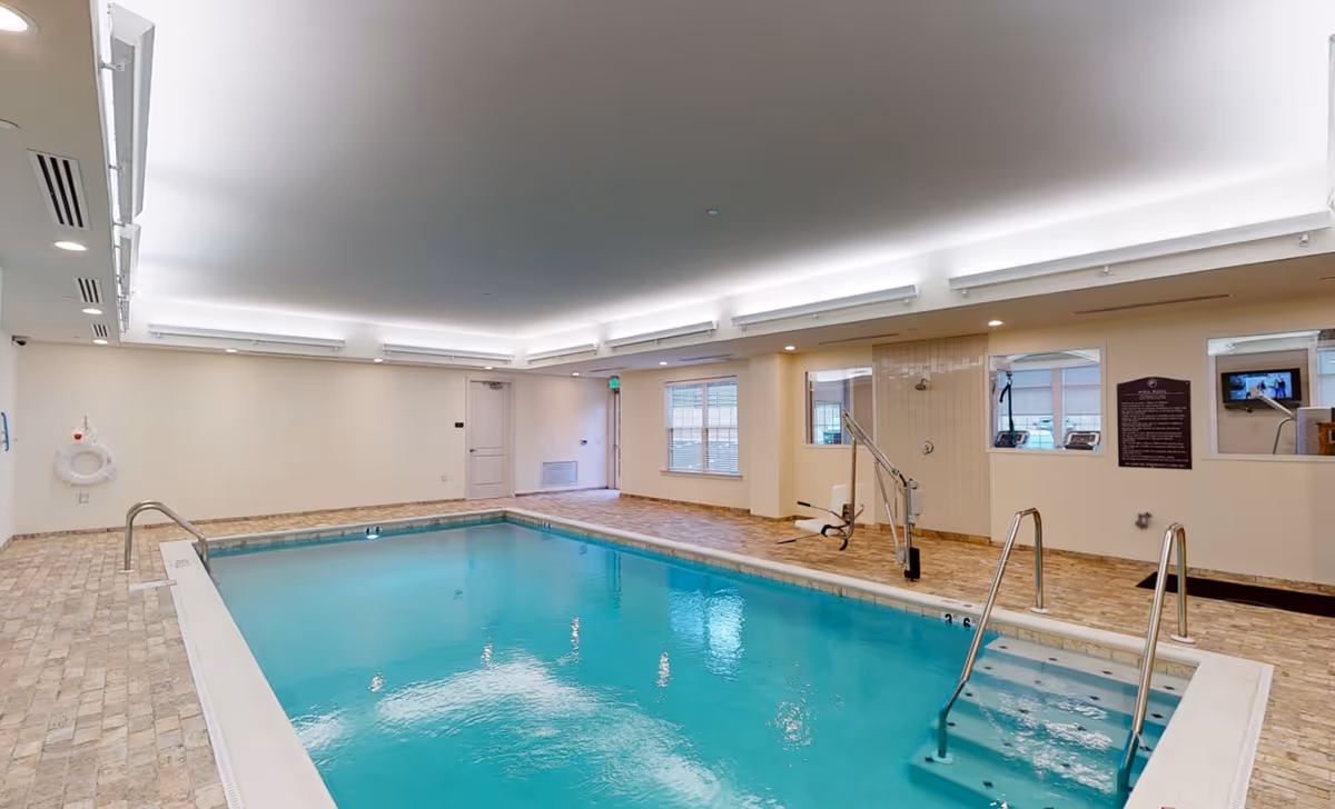 Indoor swimming pool area with clear blue water, tiled floor surrounding the pool, metal handrails, a pool lift for accessibility, and windows looking into an adjacent room with exercise equipment.