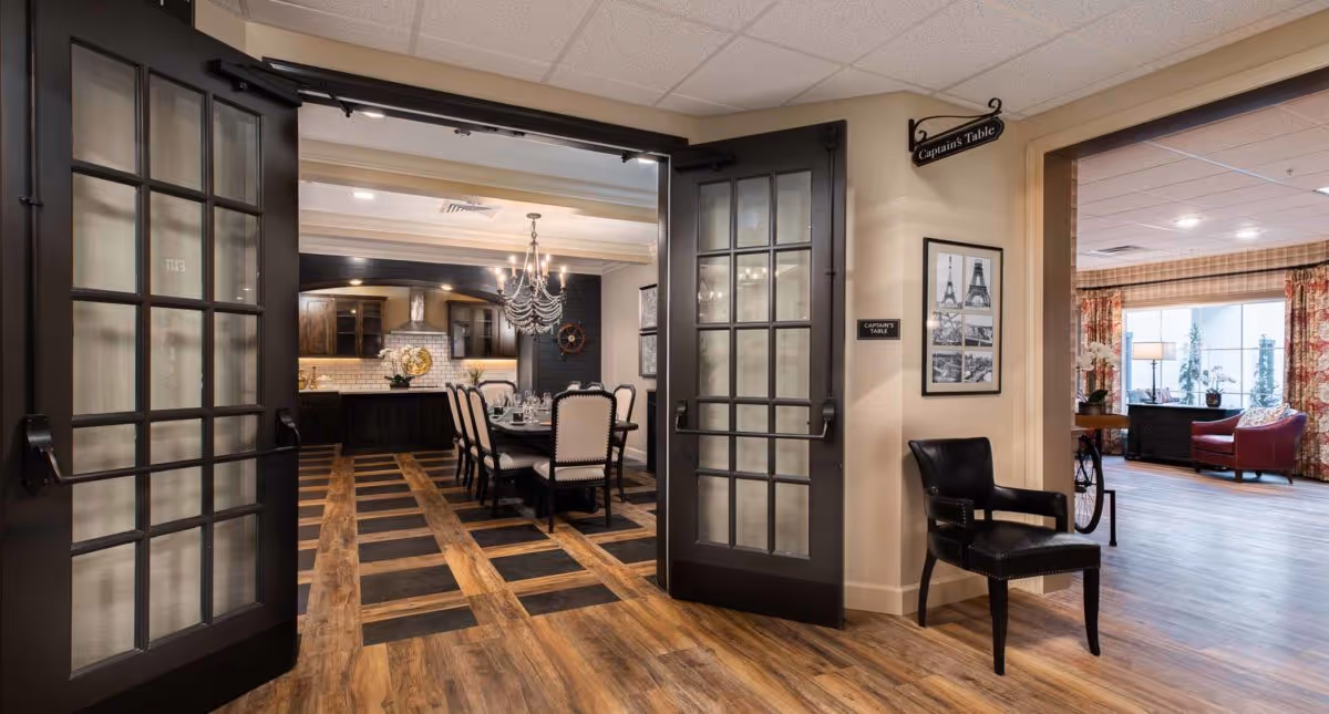 View through open double glass doors into a dining room with a long table set for a meal, surrounded by upholstered chairs. A chandelier hangs above the table. To the right, there is a black leather chair against a beige wall with framed pictures and a sign that reads 'Captain's Table.' Beyond the dining room, a living area with red chairs and large windows with floral curtains is visible.