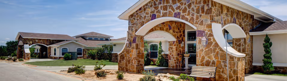 Exterior view of a senior living facility with stone archways, beige walls, and a bench near the entrance. The building is surrounded by landscaped greenery and a paved driveway.