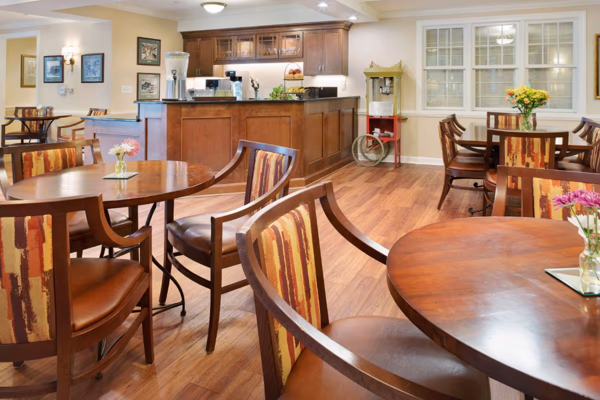 A cozy dining area in a senior living facility with round wooden tables and chairs featuring patterned upholstery. The room has wooden flooring, a counter with cabinets and beverage dispensers, a popcorn machine, and framed artwork on the walls.
