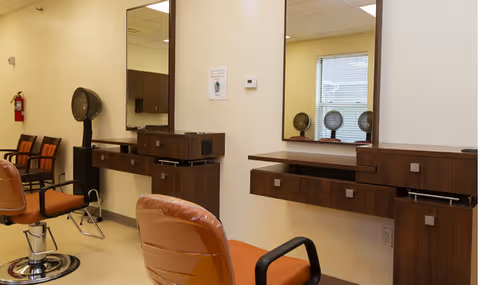 Interior view of a salon area in a senior living facility with brown salon chairs, large mirrors mounted on the wall, and hair drying equipment. There are additional chairs lined up against the wall and a fire extinguisher mounted on the wall.