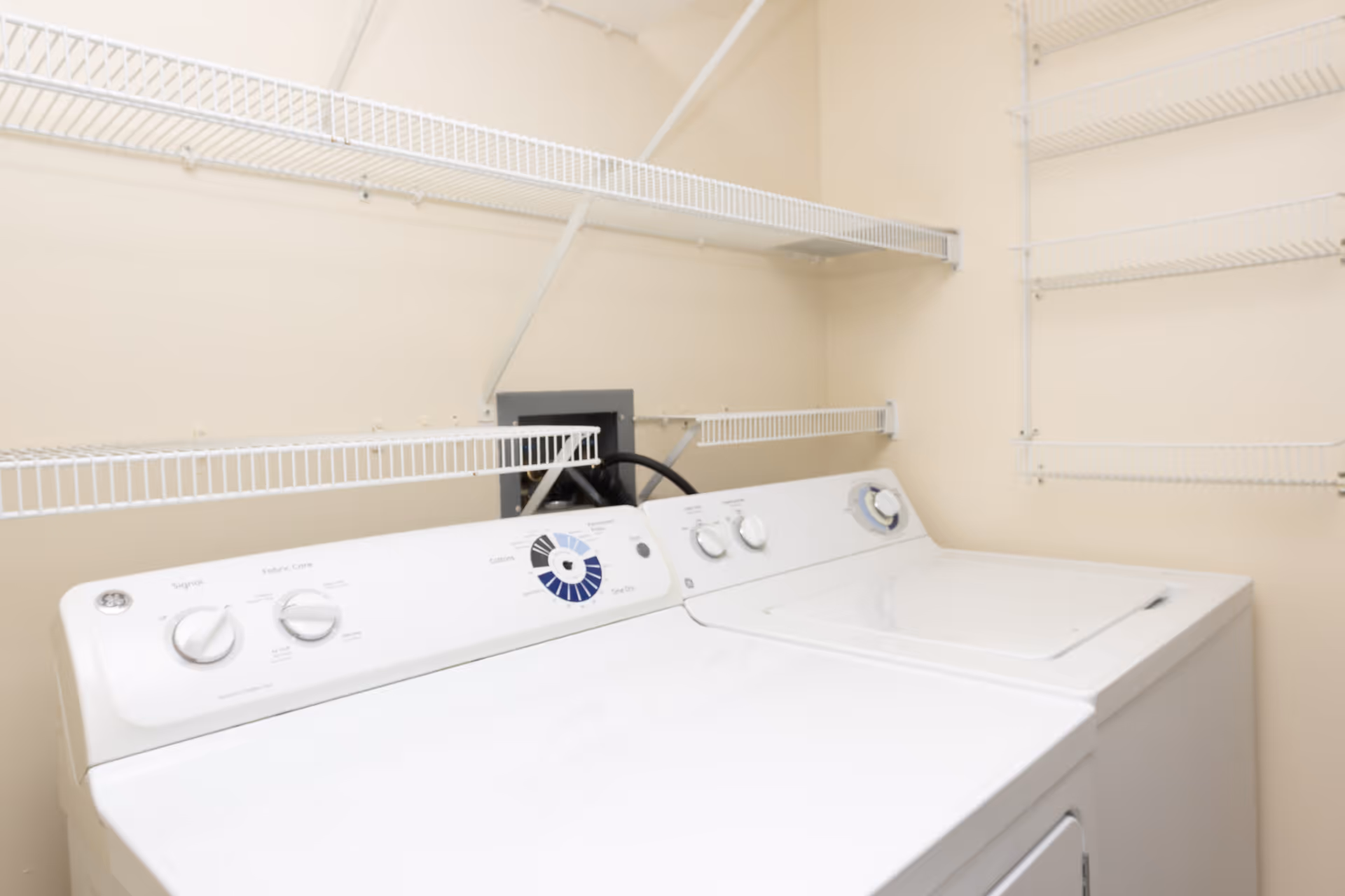 A laundry room with a washer and dryer under white wire shelving mounted on beige walls.