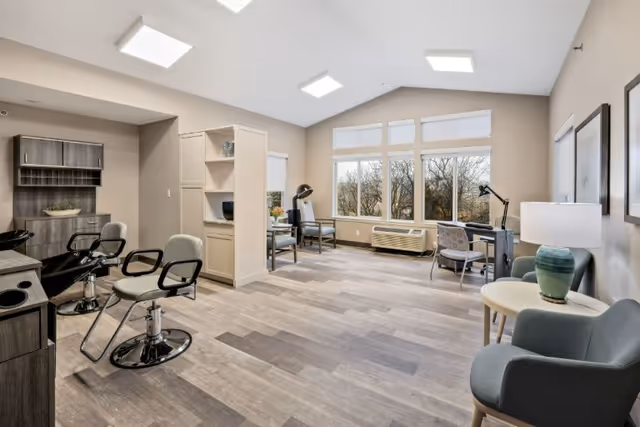 A bright, modern common room with salon chairs and workstations facing large windows in a senior living facility.