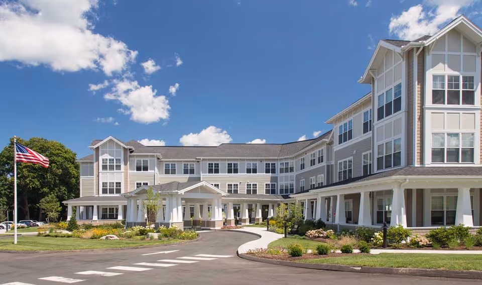 Exterior view of a large, multi-story senior living facility building under a bright blue sky with scattered clouds. The building has multiple windows, a covered entrance with white pillars, landscaped greenery, and an American flag on a flagpole in front.