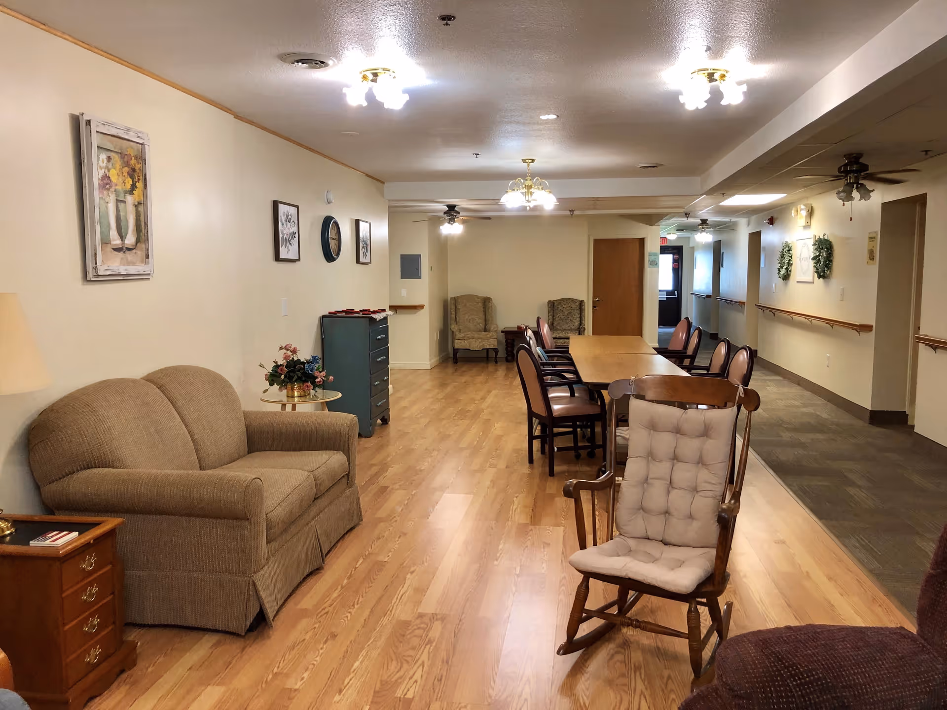 Interior view of a senior living facility common area with wooden flooring, a beige couch, a wooden rocking chair with a cushion, a long table surrounded by chairs, two armchairs in the background, wall decorations including framed pictures and a clock, and ceiling lights.
