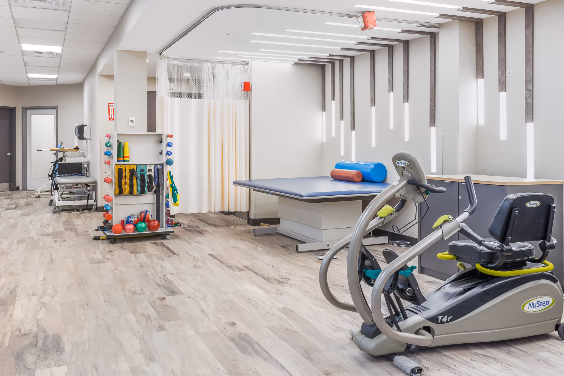 A rehabilitation room with exercise equipment including a NuStep recumbent cross trainer, a large padded therapy table with cylindrical cushions, and a rack holding various therapy balls and resistance bands. The room has light wood flooring, white walls with vertical light fixtures, and a curtain divider near the back.