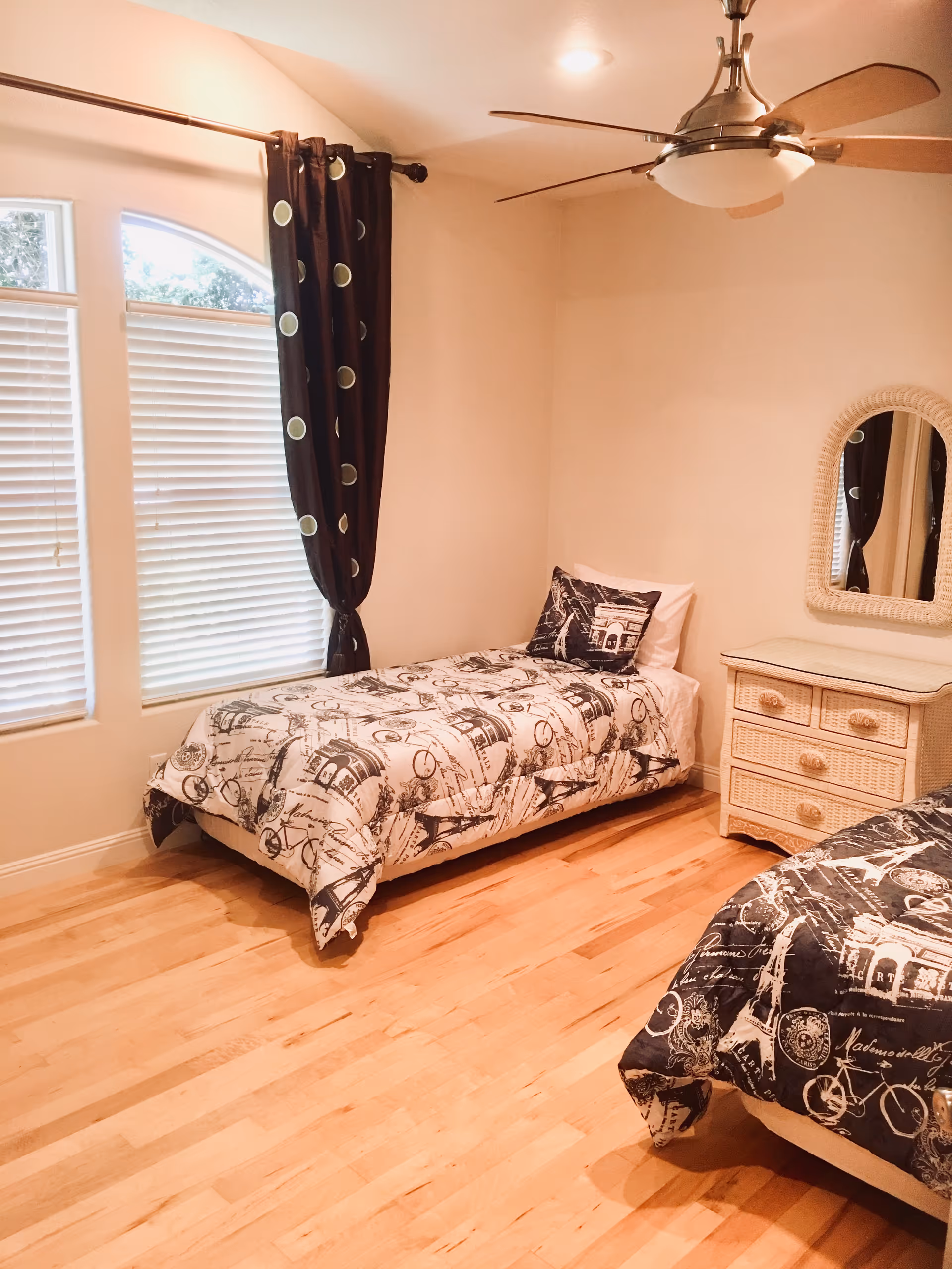 A bedroom with two twin beds featuring black and white patterned bedding. There is a wicker nightstand with drawers and a mirror above it between the beds. The room has light-colored wooden flooring, a ceiling fan with light, and a window with white blinds and dark curtains with circular patterns.