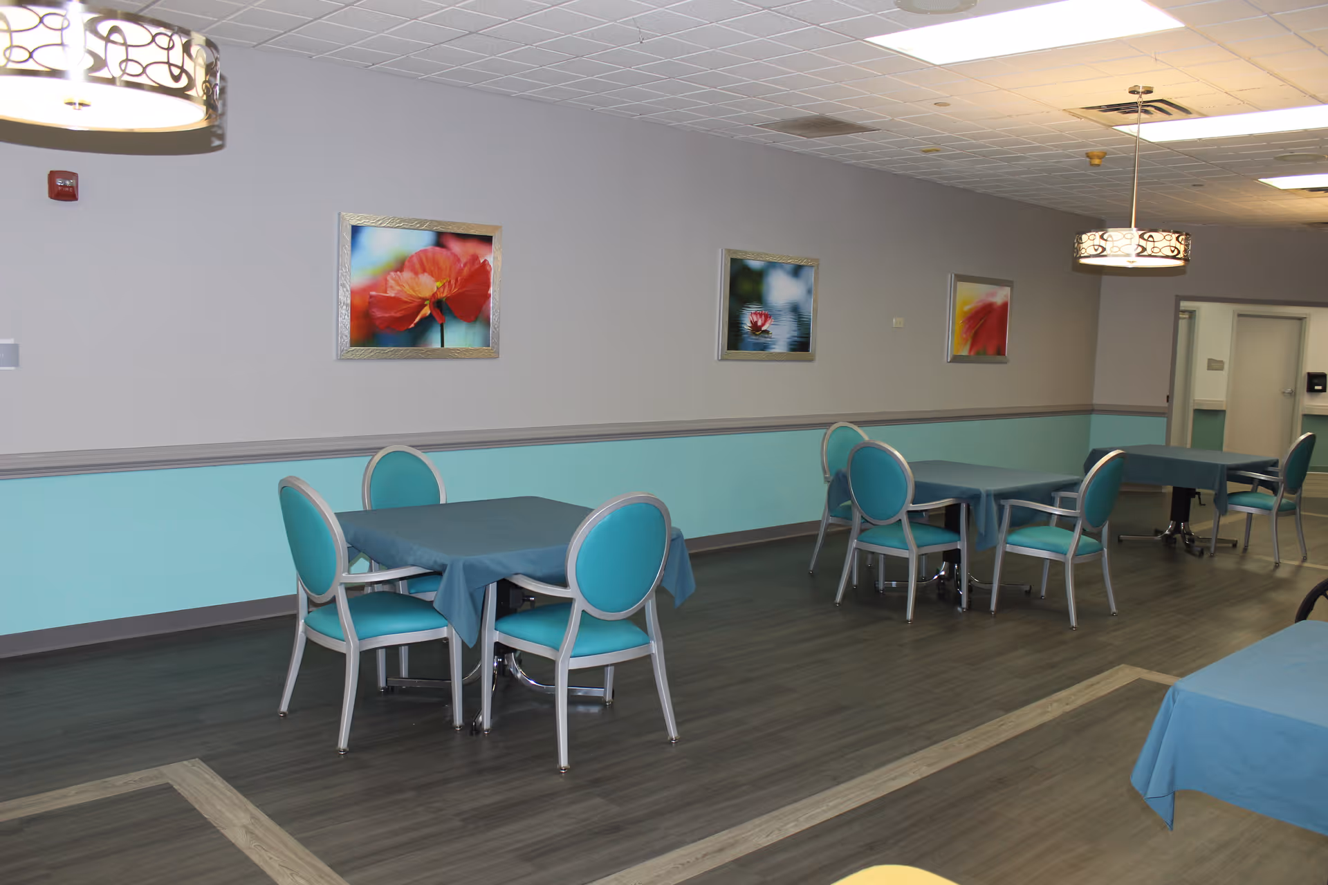 Dining room with teal-upholstered chairs and tables covered with blue tablecloths, floral wall art, and pendant lights.
