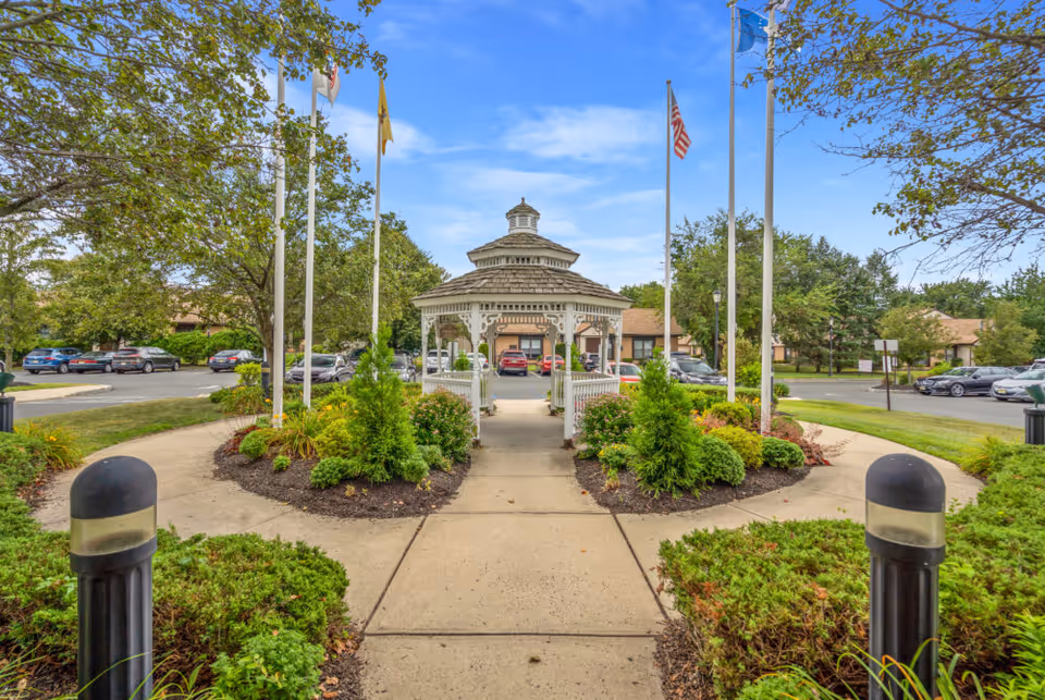 A landscaped outdoor area with a paved walkway leading to a white gazebo surrounded by green shrubs and trees. Several flagpoles with flags are positioned around the gazebo. In the background, there is a parking lot with cars and buildings under a partly cloudy blue sky.