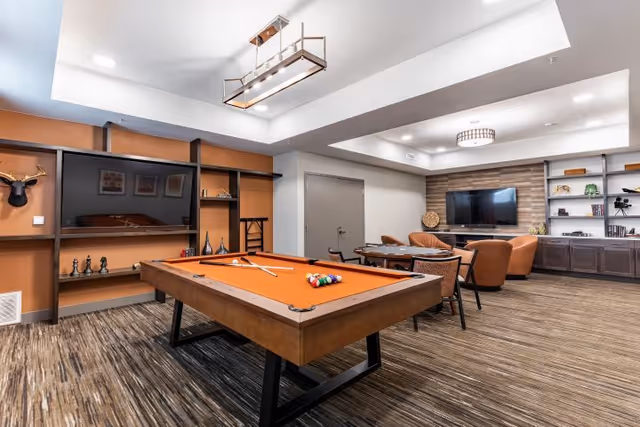 A spacious recreational room featuring an orange felt pool table with pool cues and balls arranged on it. The room has modern lighting fixtures, a large flat-screen TV mounted on a wooden accent wall, and built-in shelves with decorative items. There are several chairs and a table near the TV area, and the flooring is carpeted with a striped pattern.